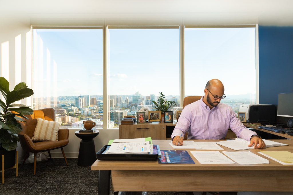 Man at desk looking over paperwork. Background includes large windows and cityscape. 